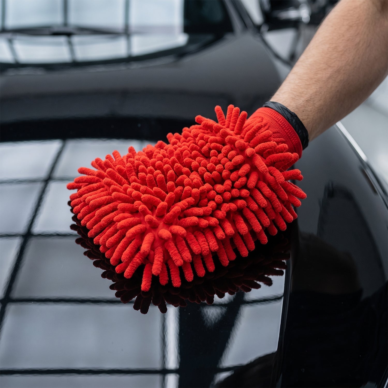 A person's hand in a red microfiber chenille wash glove cleaning the shiny black hood of a car.