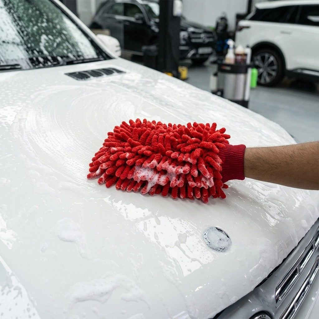 A person's hand in a red microfiber chenille wash glove covered in suds, cleaning the white hood of a car.